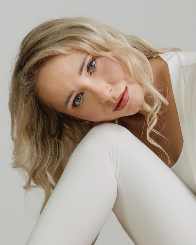 Blondynka w białym stroju opiera głowę o kolano i spogląda w obiektyw.