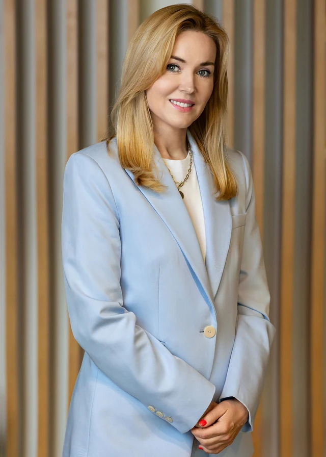 Kasia w błękitnym garniturze na tle drewnianych i szarych pionowych paneli.