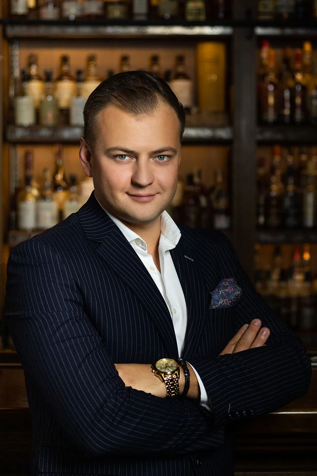 Mężczyzna w granatowym garniturze w prążki, ze skrzyżowanymi ramionami, na tle eleganckiego baru.
