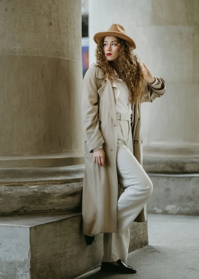 Julia w beżowym płaszczu i kapeluszu, w jasnym stroju przy kamiennych kolumnach.